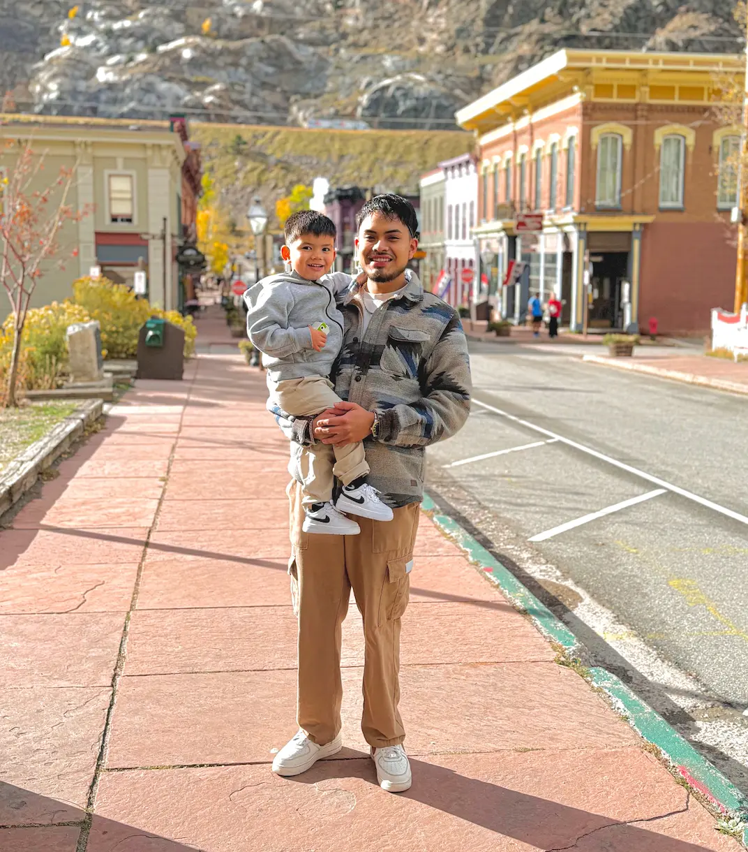 Edgar, owner of Chino's Remodel & Build, holding his young son on a brick sidewalk in a historic Colorado mountain town