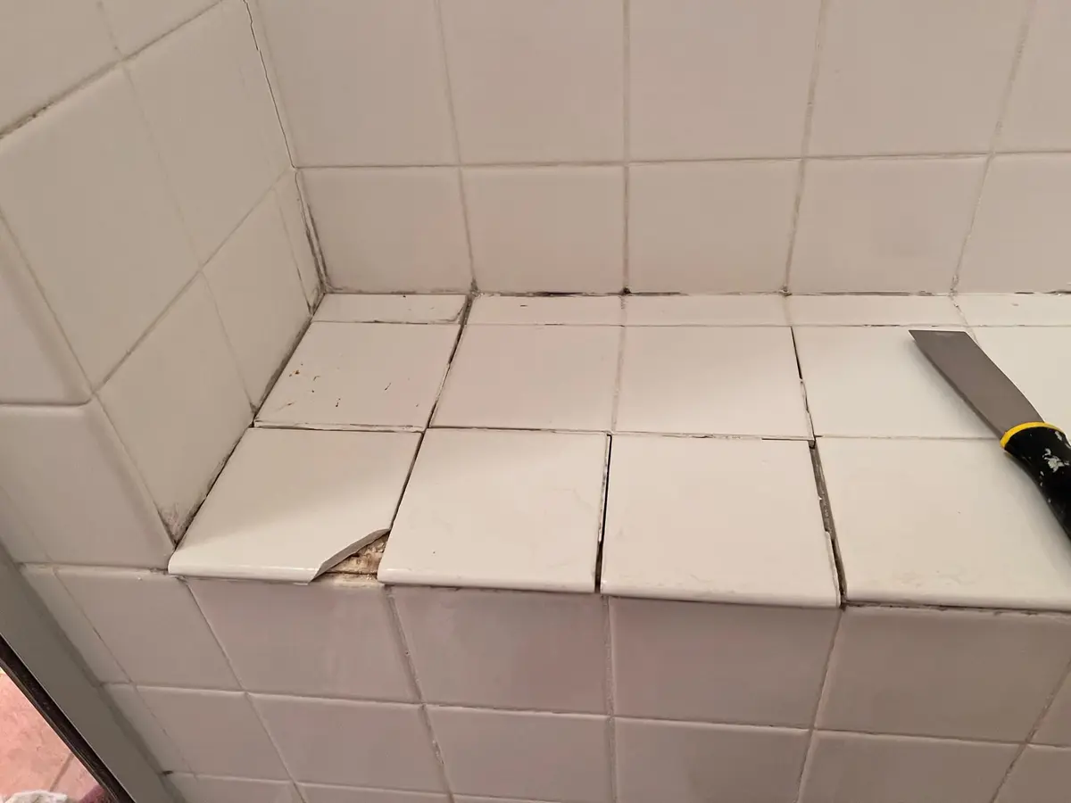 Tiled shower bench with cracked and loose tiles and a scraper resting on top