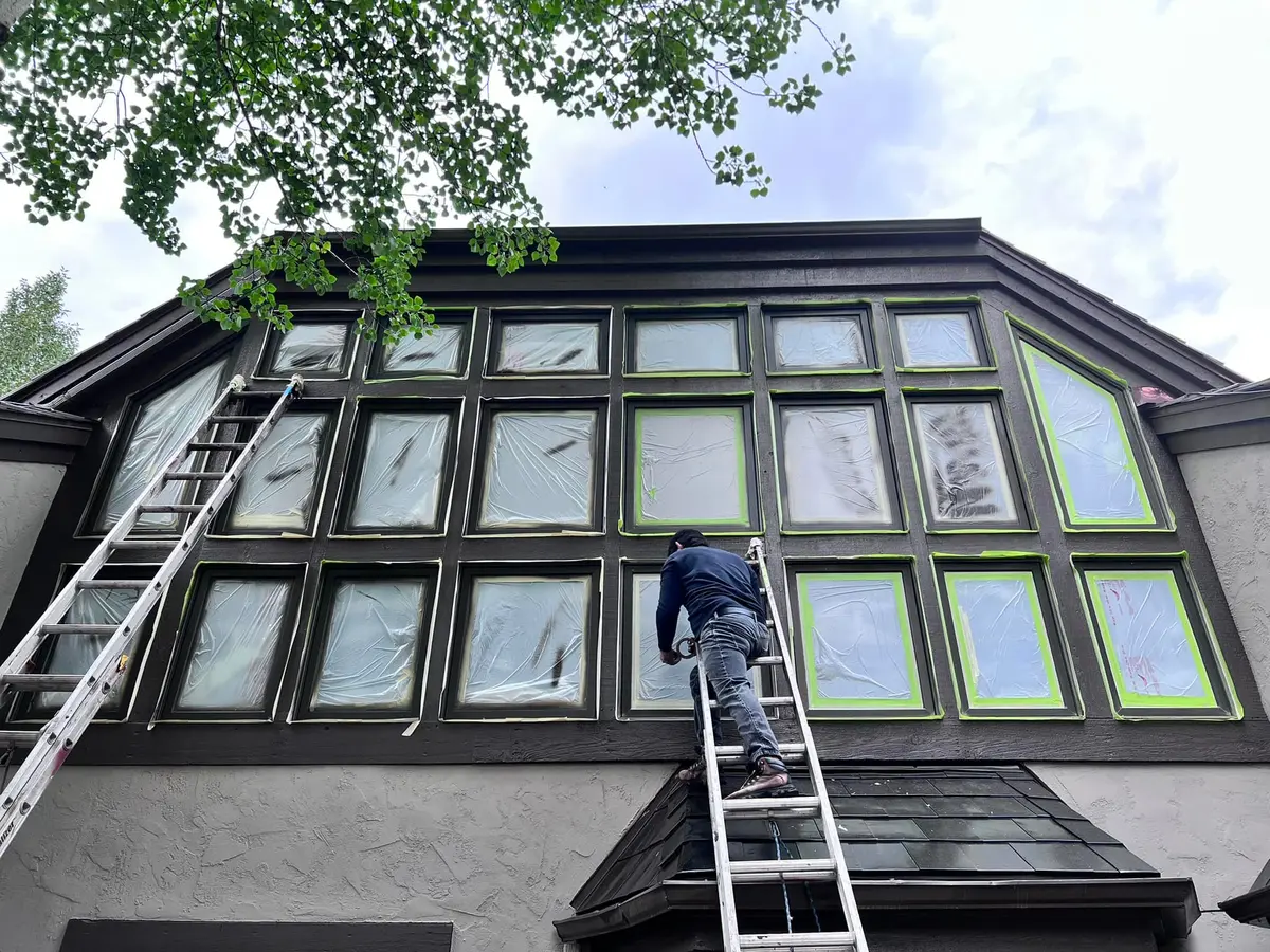 Edgar on a ladder restoring the frames of a large multi-pane window wall on a stucco and stone mountain home.